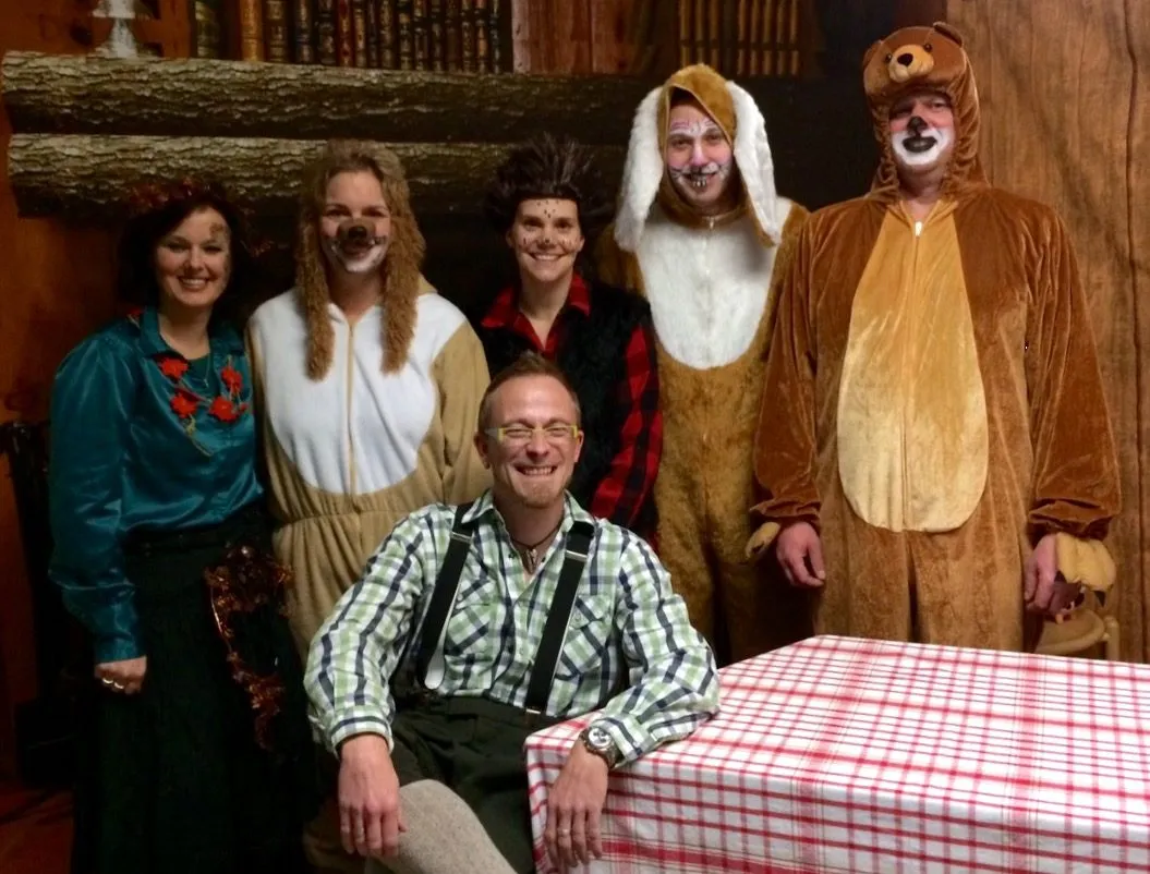 Gruppenfoto des Ensembles von „Winter im Wald“ in Kostüm — Förster, Hase, Igel, Fuchs, Bär und eine Figur im grünen Kleid vor einer Holzkulisse mit gedecktem Tisch.