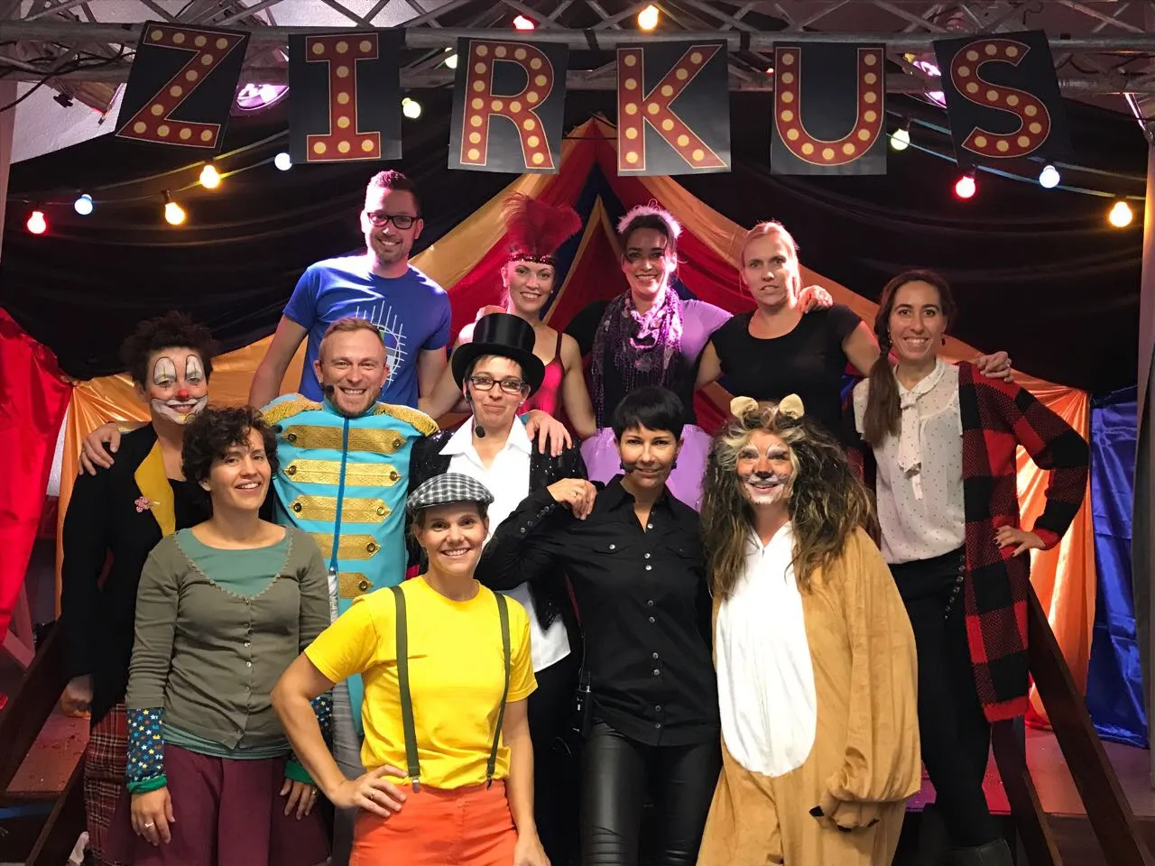 Gruppenfoto des Ensembles von „Nimm Platz im Zirkus Karamboli“ in Kostüm — Clown, Löwe, Zirkusdirektor und weitere Figuren unter dem Schriftzug „Zirkus“.