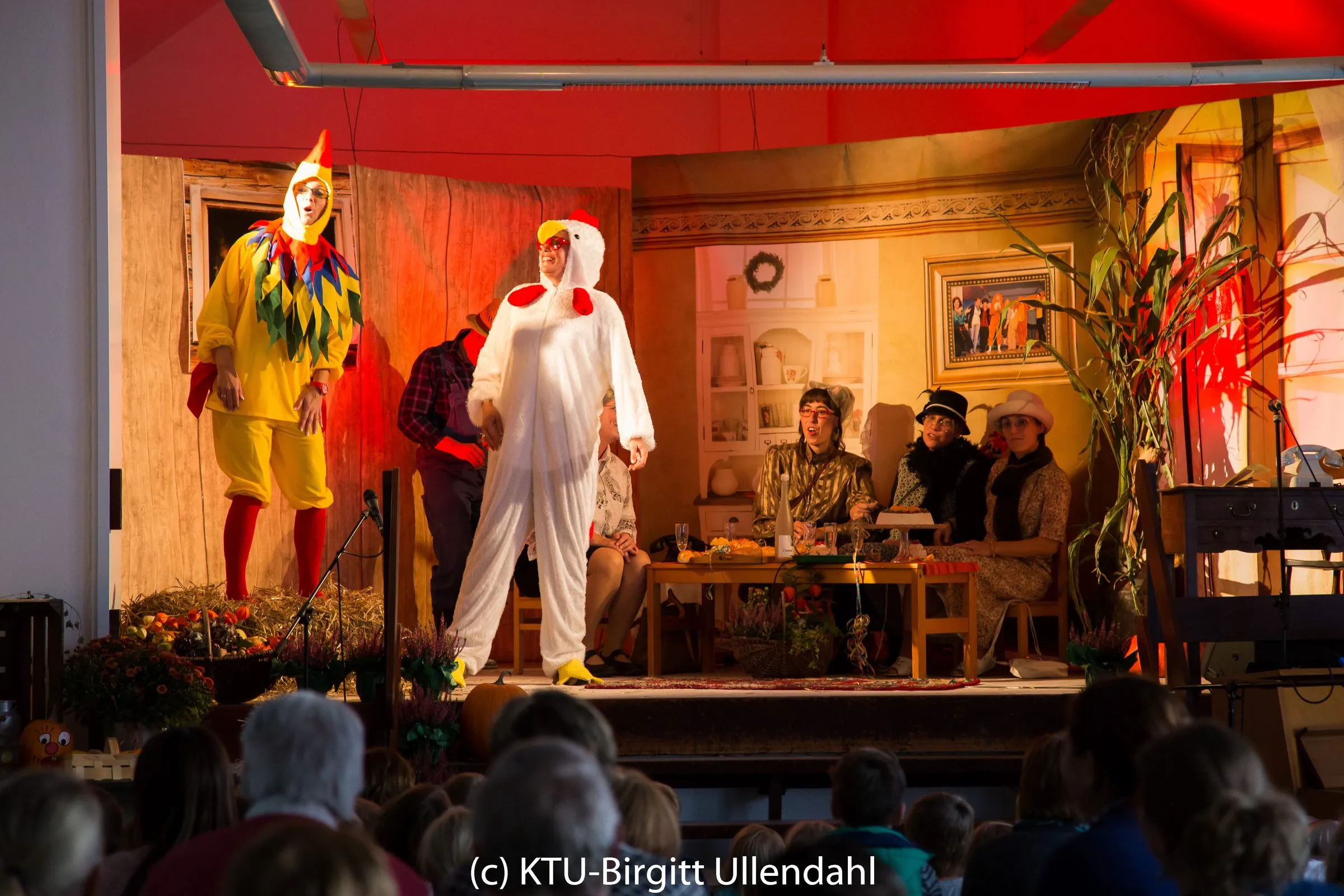 Hahn, Huhn und Kuh im Kostüm applaudieren neben Bauer Anton vor der Damenrunde am Tisch.