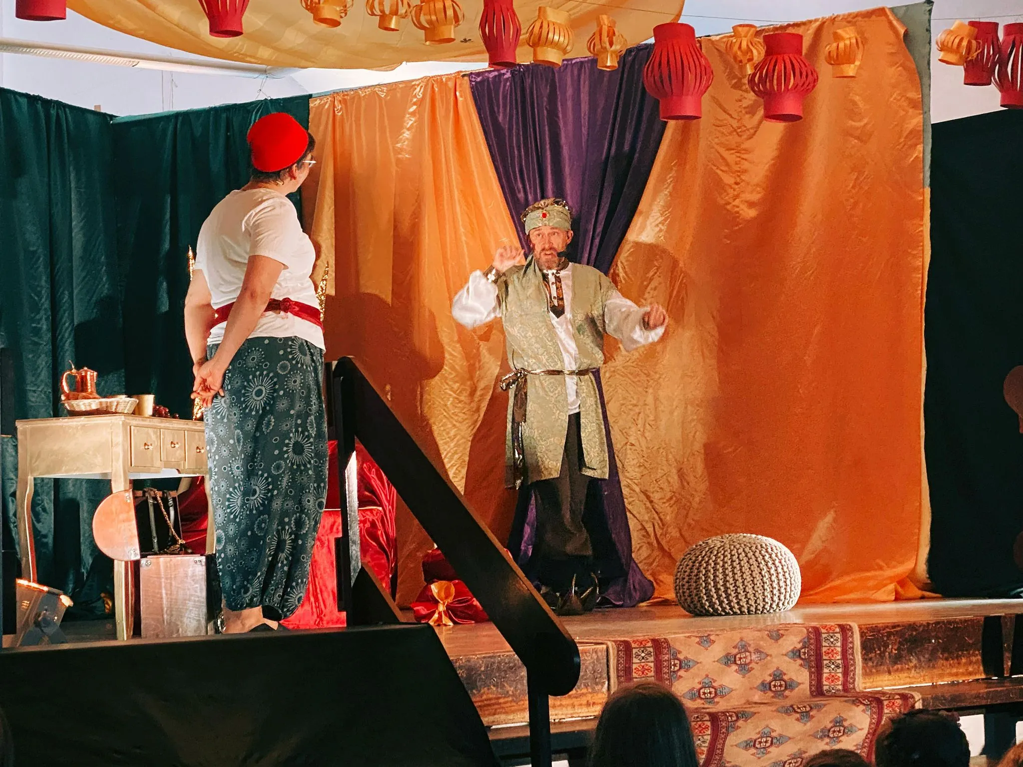 Eine Figur mit rotem Fes im Gespräch mit dem Sultan vor lila und goldenen Vorhängen.