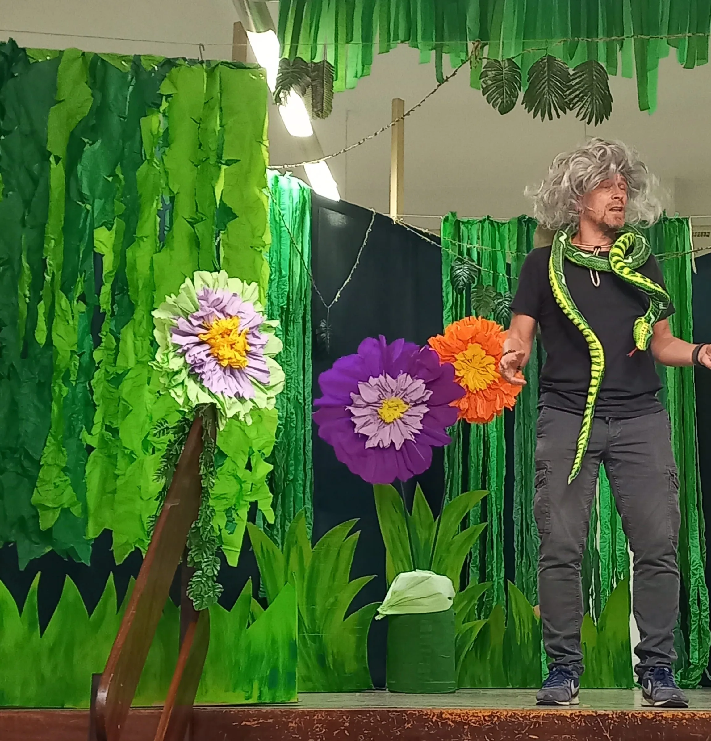 Die Figur mit grauer Perücke und Schlangenrequisite vor großen Papierblumen.