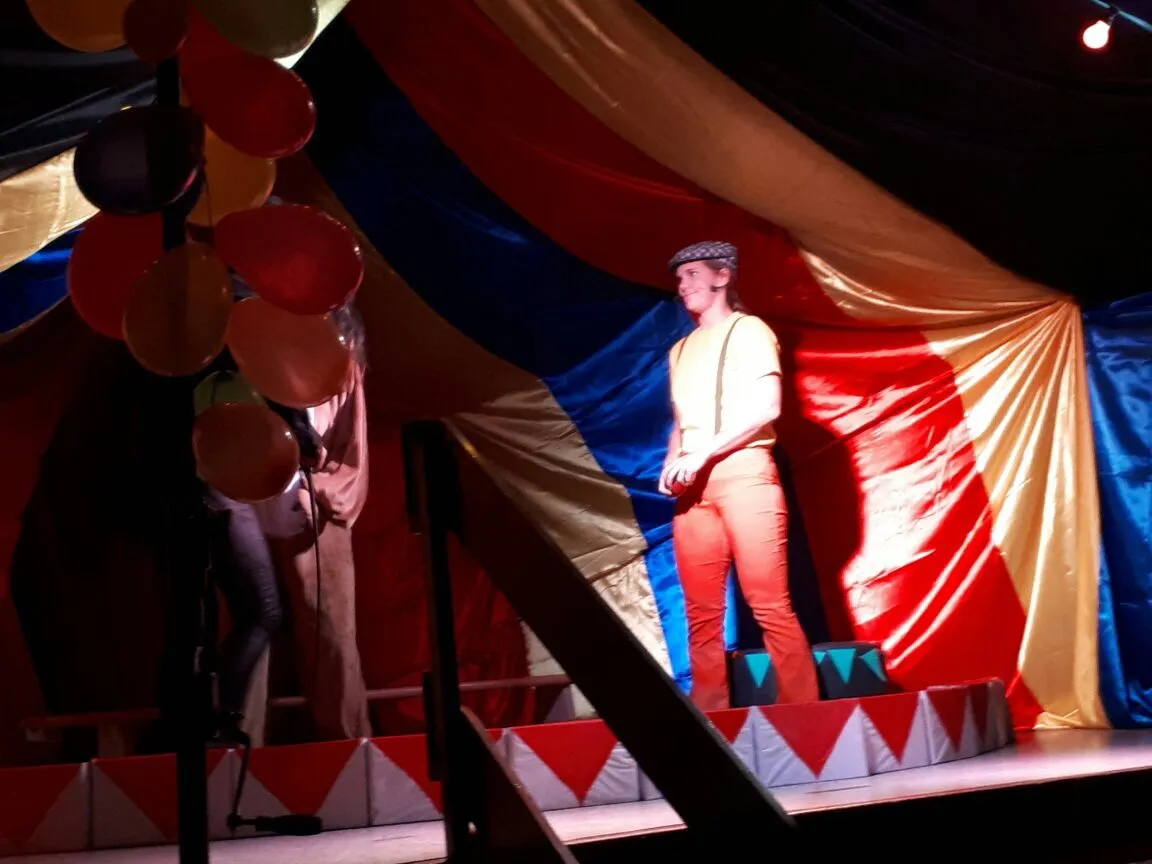 Jongleur Jojo mit Schiebermütze posiert vor der bunten Zeltkulisse.