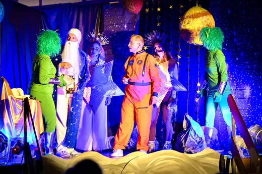 Edwin im orangefarbenen Astronautenanzug im Gespräch mit dem Mann im Mond, zwei Sternen und zwei grünen Außerirdischen vor einer blau beleuchteten Sternenkulisse mit Planeten.