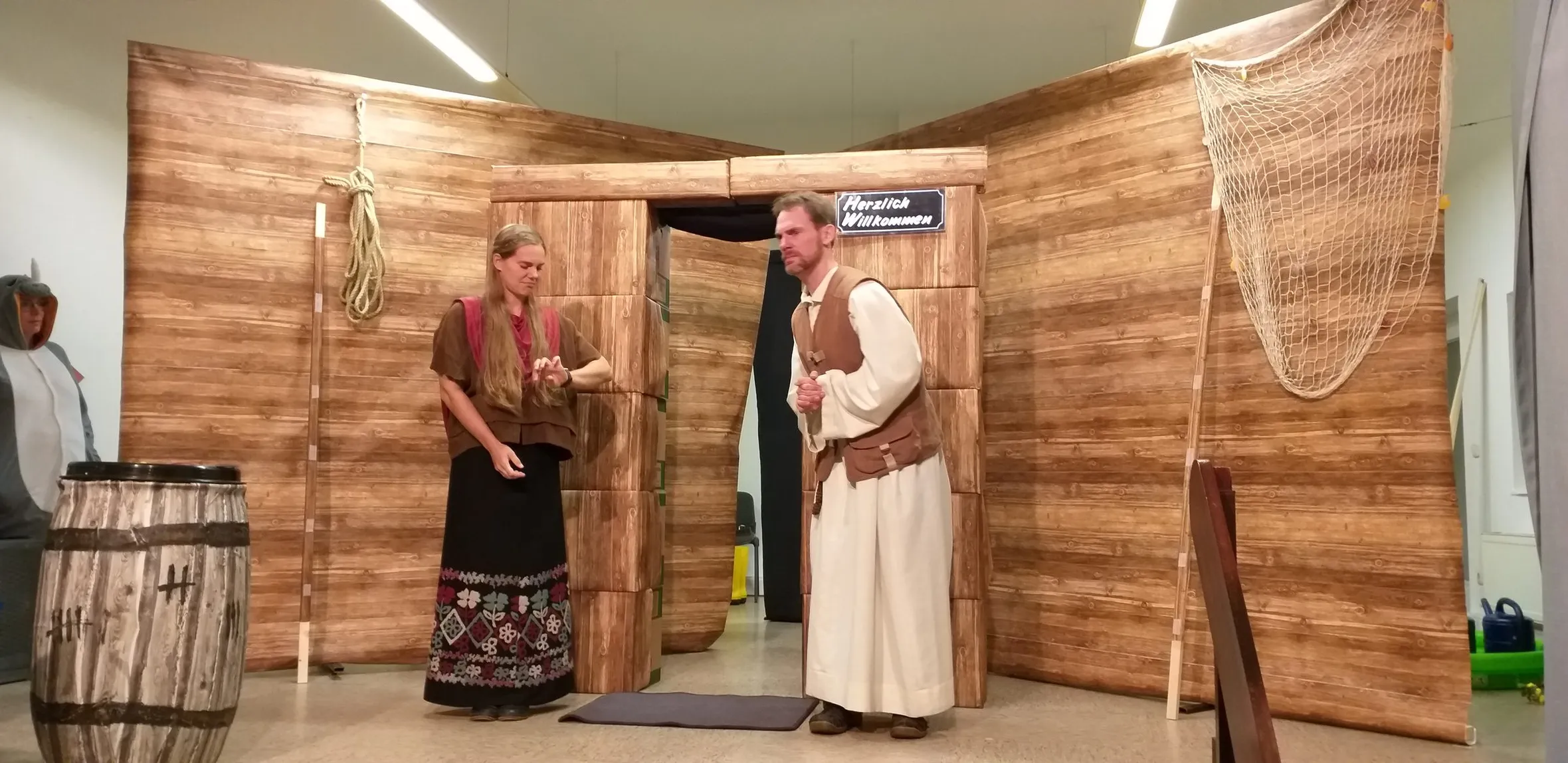 Zwei Figuren im Gespräch vor der Bühnenkulisse einer Arche aus Holzplanken mit Tau, Fischernetz und dem Schild „Herzlich Willkommen“.