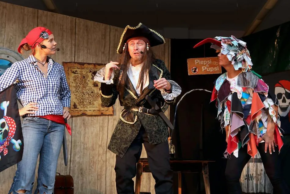 Käpt'n Klaas im Gespräch mit zwei weiteren Piratenfiguren vor der Bühnenkulisse, im Hintergrund eine Schatzkarte und das Schild „Piepsi & Pupsi".